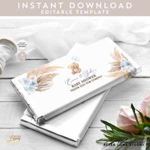 May include: A white chocolate bar wrapper with a blue and brown design featuring a teddy bear holding balloons, pampas grass, and flowers. The text on the wrapper reads "Emma & John's Baby Shower Thank You for Coming!"