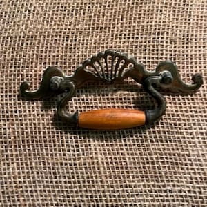 May include: A drawer pull featuring a dark metallic backplate with a decorative shell motif and curved arms. A smooth, cylindrical wooden handle with a warm brown finish is suspended between the two metal arms of the hardware piece.