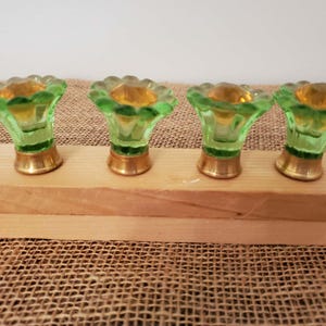 May include: Four vintage green glass candle holders with gold-colored bases and tops. The candle holders are flower-shaped and sit on a wooden plank. The glass has a slight yellow tint.