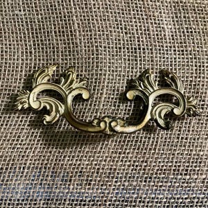 Puede incluir: Tirador de cajón de latón ornamentado con un asa curvada y detalles decorativos florales y de volutas. El herraje tiene una estética vintage y está colocado sobre un fondo neutro texturizado.