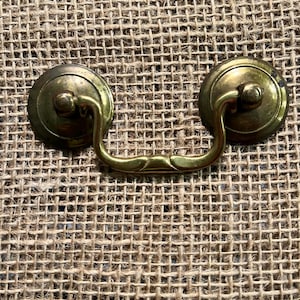 May include: Close-up of a vintage brass drawer pull with a decorative handle. The pull features two circular backplates and a curved handle with a floral design. The hardware is set against a textured burlap background.