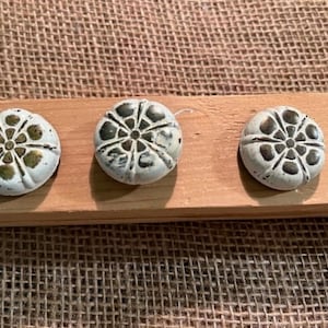 May include: Three round buttons with a white finish and a recessed floral pattern featuring dark metallic accents. Each button has a scalloped edge and a symmetrical design with a central point radiating outward into eight distinct petal shapes.