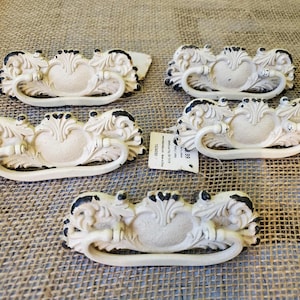May include: Five antique-style drawer pulls with a cream-colored finish and black accents. Each pull features an ornate design with a central heart-shaped element and a curved handle. The pulls are arranged on a textured, tan surface.