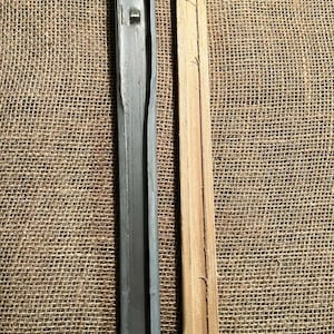 May include: Two long, narrow pieces. One is a weathered, gray metal channel with a rectangular cross-section. The other is a light-colored wooden slat with a groove and a hole. Both are positioned vertically against a textured, tan background.