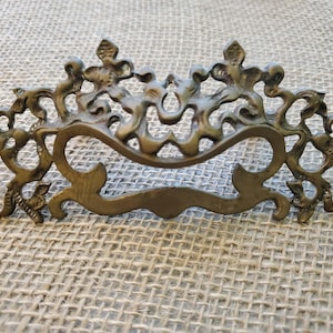 May include: Ornate antique brass drawer pull with a scrolling design. The decorative hardware features a curved handle and intricate floral and foliate details. The aged brass finish adds a vintage aesthetic.