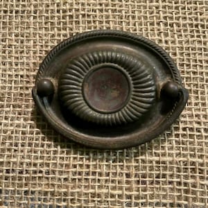 May include: Oval-shaped antique bronze drawer pull with a central circular design and a rope-like border. Two small, round knobs are on either side of the pull. The hardware is set against a textured, tan background.