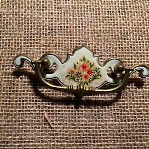May include: A vintage drawer pull with a floral design. The pull has a brass handle and a decorative center piece with a light blue background and a floral pattern in red, pink, and yellow.