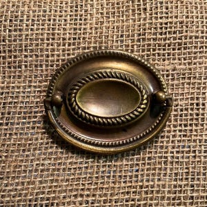 May include: An oval-shaped brass drawer pull featuring a central recessed handle surrounded by a decorative rope-style border and a beaded outer edge. The metal hardware has a dark, aged patina with visible highlights on the raised surfaces.