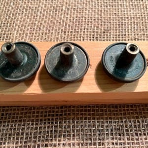 Puede incluir: Tres botones de metal de color oscuro con un dise&ntilde;o circular y un poste central sobresaliente. Cada bot&oacute;n presenta un peque&ntilde;o orificio en la parte superior del poste y texto visible grabado alrededor de la base, con un acabado met&aacute;lico oscuro.