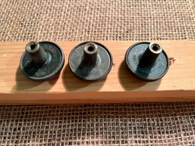 Puede incluir: Tres botones de metal de color oscuro con un dise&ntilde;o circular y un poste central sobresaliente. Cada bot&oacute;n presenta un peque&ntilde;o orificio en la parte superior del poste y texto visible grabado alrededor de la base, con un acabado met&aacute;lico oscuro.