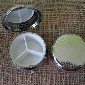 May include: A silver pill box with a hinged lid and three compartments. The pill box is open and the compartments are visible.
