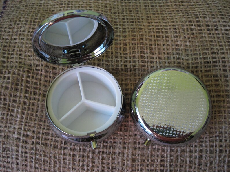 May include: A silver pill box with a hinged lid and three compartments. The pill box is open and the compartments are visible.