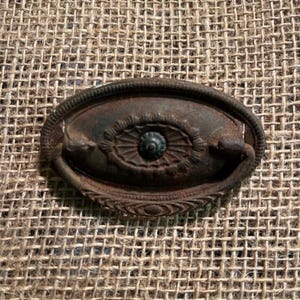 Puede incluir: Un tirador de cajón ovalado de metal con un acabado oscuro y desgastado. El herraje presenta un medallón circular central rodeado de intrincados patrones radiales en relieve y un asa curva articulada sujeta a los lados de la placa.