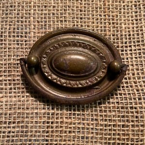 May include: Antique brass drawer pull with an oval center and decorative detailing. The pull has two small, round knobs on either side and a dark, aged finish. The hardware is likely for a cabinet or drawer.