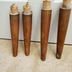 Pode incluir: Quatro pernas de móveis de madeira vintage com um design cónico. As pernas são de uma cor castanha quente, mostrando sinais de desgaste e idade. Cada perna tem um topo de madeira de cor clara, provavelmente para fixar a um móvel.