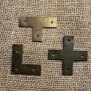 May include: Three antique brass-toned metal brackets in different shapes: a T-shape, an L-shape, and a cross-shape. Each bracket has pre-drilled holes for mounting. The brackets are displayed on a textured, neutral-colored surface.