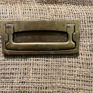 May include: A rectangular antique brass drawer pull with a recessed handle. The pull has a slightly curved top and is mounted on a textured, tan burlap surface. The hardware is likely for a cabinet or drawer.