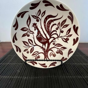 May include: A white ceramic plate with a brown rooster and tree design. The plate features a detailed illustration of a rooster perched on a tree branch with leaves and berries. Decorative accents surround the rim.