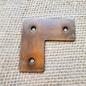 May include: A bronze-colored, L-shaped metal bracket with three circular holes for screws. The bracket has a slightly aged appearance, with variations in color and texture. The item is likely used for joining or reinforcing corners in furniture or other woodworking projects.