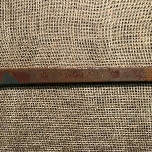 May include: A rectangular, rusty-brown metal object with a slightly textured surface. The object has a faded, weathered appearance with patches of blue-gray paint on the ends. The item is set against a neutral, textured background.
