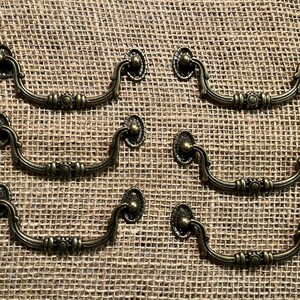 May include: Six antique bronze drawer pulls with a decorative floral design. The pulls have a curved handle and are mounted on a textured background. These vintage-style pulls are ideal for furniture restoration projects.