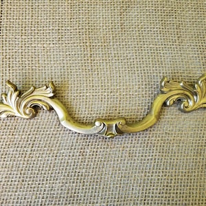 May include: Ornate brass drawer pull with a curved handle and decorative floral scrollwork on each end. The hardware has a vintage aesthetic and is likely for a cabinet or dresser.