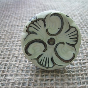 May include: A round, off-white ceramic cabinet knob with a floral design. The design features a central circle with radiating lines and curved elements, all outlined in dark brown. The knob is set against a textured, neutral-toned background.