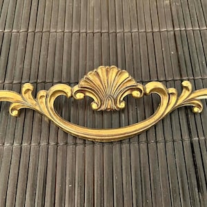 May include: Ornate gold-colored drawer pull with a shell-shaped center and scrolling leaf designs on each end. The hardware is made of metal and has a vintage aesthetic. The pull is approximately 8 inches long.