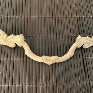 Pode incluir: Uma puxador de móvel de madeira curvado, bege claro, com desenhos florais e de folhas esculpidos. O puxador tem um detalhe floral central e mede aproximadamente 25 cm de comprimento.