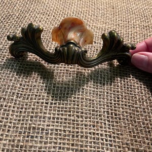 May include: A drawer pull featuring an ornate, curved brass-toned metal base with intricate scrollwork and a central, smooth, translucent, marbled stone-like insert displaying swirls of cream, tan, and reddish-brown colors.