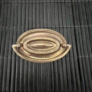 Op de afbeelding: Een messingkleurige ovale ladegreep met een centraal verzonken ontwerp en gebogen handgrepen. Deze meubelbeslag heeft een vintage uitstraling. De greep staat tegen een donkere, getextureerde achtergrond.