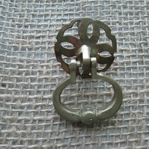 May include: Antique brass drawer pull with a decorative floral design and a ring handle. The hardware is mounted on a textured, woven fabric background. This vintage-style pull is suitable for cabinets and drawers.