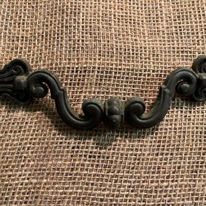 May include: A dark bronze-toned metal drawer pull featuring an ornate, curved design with symmetrical scrollwork and a central decorative element. The handle has a weathered, matte finish with intricate detailing on both ends and a smooth, arched center.