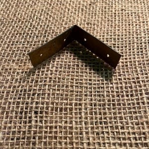 May include: A dark brown metal corner brace featuring an L-shaped design with two flat arms meeting at a ninety-degree angle. Each arm of the metal bracket contains two small circular pre-drilled holes intended for mounting screws or fasteners.
