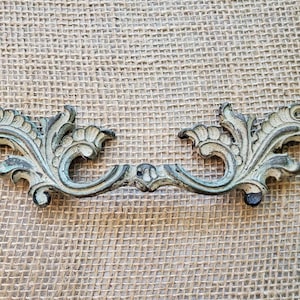 May include: An antique-style drawer pull with an ornate, floral design. The metal hardware features a distressed, aged appearance with a mix of green, brown, and cream colors. The pull is approximately 6 inches long.