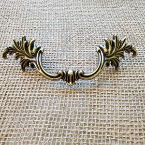 Könnte beinhalten: Verzierter antiker Schubladengriff aus Messing mit gebogenem Griff und dekorativen Blattakzenten. Die Hardware besteht aus Metall und ist für Möbel konzipiert. Der Griff ist etwa 15 cm lang.