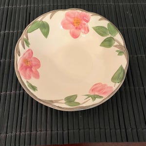 May include: A round ceramic bowl with a scalloped edge and a cream-colored base. The interior features a hand-painted floral pattern with three pink flowers, green leaves, and brown stems. The rim of the bowl is finished with a thin, dark gray line.