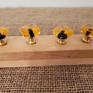 May include: Four decorative cabinet knobs with a unique design. Each knob features a clear, amber-colored glass top with a black accent, mounted on a brass base. The knobs are arranged on a wooden plank, showcasing their design and potential use.