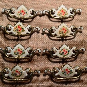 May include: Six vintage drawer pulls with a floral design. Each pull features a white enamel center with a bouquet of red, pink, and green flowers. The pulls have a gold-toned metal frame and curved handles, perfect for antique furniture restoration.