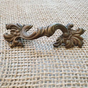May include: An antique-style bronze drawer pull with a decorative, symmetrical design. The hardware features detailed floral and scrollwork patterns, adding a vintage aesthetic. The pull is likely made of metal and is suitable for furniture restoration or decorative projects.