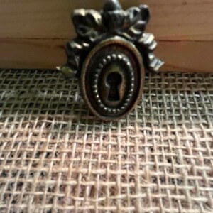 May include: A metallic keyhole cover plate featuring an oval center with a beaded border and a decorative bow-shaped top. The hardware has a dark, aged bronze finish with visible highlights on the raised edges and a central vertical keyhole slot.