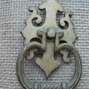Peut inclure: Poignée de tiroir en laiton antique avec une plaque arrière décorative et une poignée en boucle. La poignée présente un motif de corde torsadée. La quincaillerie a une patine vieillie, suggérant une utilisation vintage. Pour meubles.