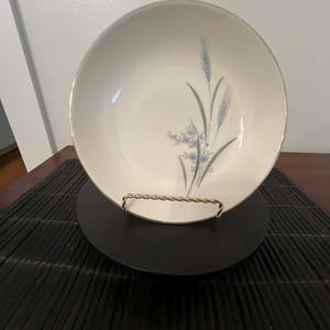May include: A white ceramic bowl with a silver rim, featuring a delicate floral design in blue and gray. The bowl is displayed on a black stand with a gold-colored wire frame, set against a dark background.