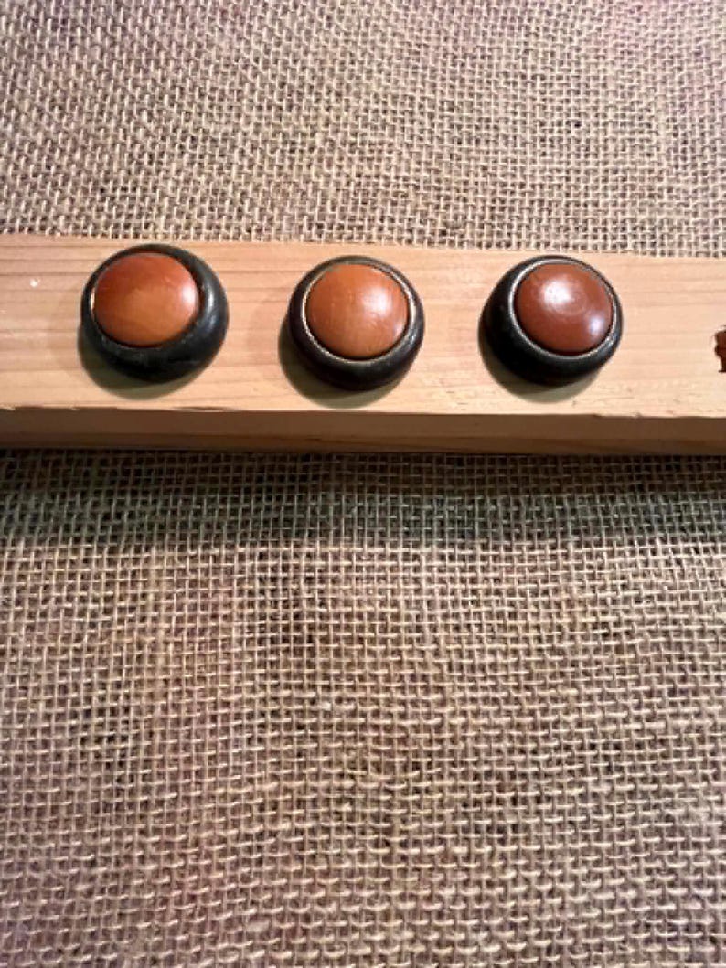 Puede incluir: Tres botones circulares con centros lisos de color marr&oacute;n claro y bordes exteriores redondeados de color marr&oacute;n oscuro. Los botones est&aacute;n montados en una fila horizontal sobre un bloque de madera rectangular de color claro, contrastando los tonos.