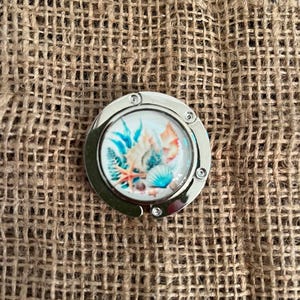 May include: A circular silver-toned metal bag hanger featuring a central decorative insert with a colorful illustration of various seashells, a starfish, and aquatic plants against a white background, secured by small rivets along the outer rim.