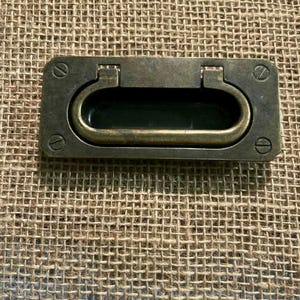 May include: A rectangular, antique brass-toned recessed pull with an oval handle. The pull is secured with four screws at each corner. The handle is slightly raised and has a dark interior.