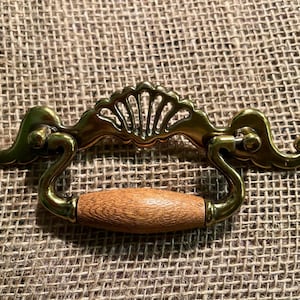 Puede incluir: Un tirador de cajón decorativo de latón con un mango de madera. El latón tiene un diseño ornamentado con un detalle en forma de abanico en el centro. El mango es de color marrón claro.