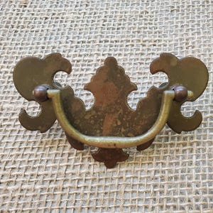 May include: Antique brass drawer pull with a decorative design. The pull features a curved handle and ornate backplates with a distressed finish. The hardware includes two small, round, brown accents on each side of the handle.