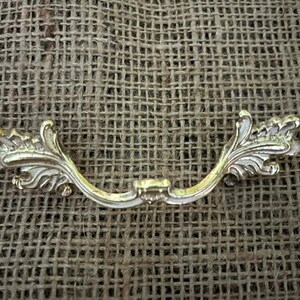 Puede incluir: Un tirador de cajón dorado y ornamentado con un diseño curvo. El tirador presenta un punto de montaje central y adornos decorativos en forma de hoja en cada extremo. El herraje se encuentra sobre un fondo marrón texturizado.