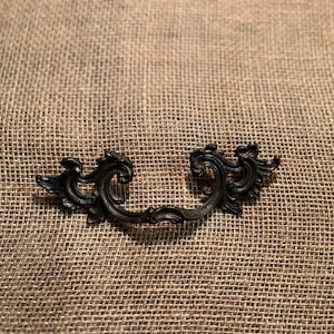 Könnte beinhalten: Verzierter schwarzer Metall-Schubladengriff mit einem dekorativen, gerollten Design. Der Griff ist auf einer strukturierten, beigefarbenen Jutefläche zentriert. Diese Hardware im Vintage-Stil.